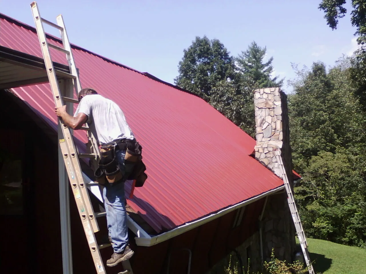 Roofer on a ladder beside a red metal roof and stone chimney — Soffit & Fascia Repair by Roofing Brothers in Manitowoc